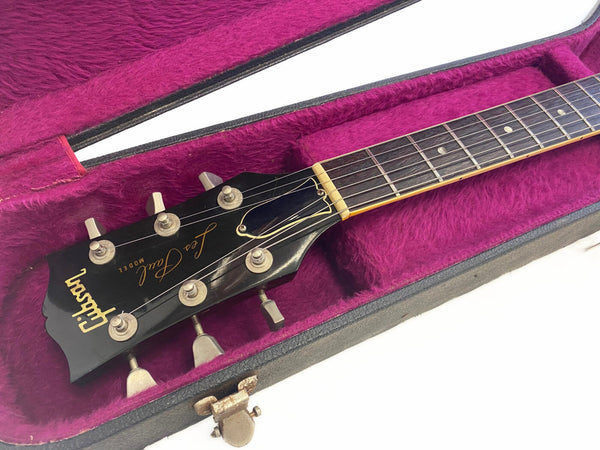 Close-up of Gibson Les Paul guitar headstock and fretboard inside purple plush-lined black hard case showing tuning pegs and nut