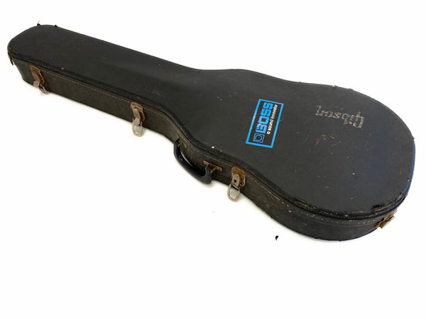 Vintage black Gibson hard guitar case with worn leather latches and BOSS sticker on top, rectangular handle, and scuffed exterior surface on white background.