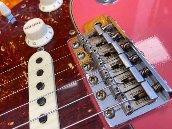 Close-up of red electric guitar bridge, Fender stamped saddles, white volume knob, and tortoiseshell pickguard with single-coil pickup and steel strings