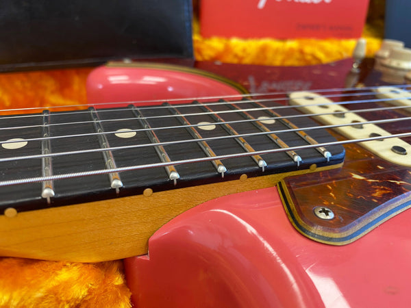 Close-up view of electric guitar neck and fretboard with metal strings, tortoiseshell pickguard, and coral pink body resting on orange plush lining.