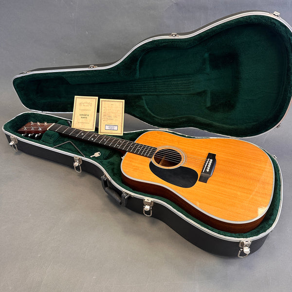 Acoustic guitar with natural wood finish in black hard case with green plush interior, warranty cards, and guitar key on gray background