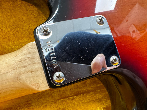 Close-up of electric guitar neck plate with serial number V071231 on sunburst guitar body and natural wood neck against a yellow fabric background