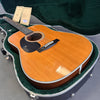 Acoustic guitar with natural wood finish and black pickguard resting in green velvet-lined hard case with warranty cards and guitar key.