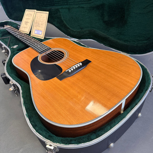 Acoustic guitar with natural wood finish and black pickguard resting in green velvet-lined hard case with warranty cards and guitar key.