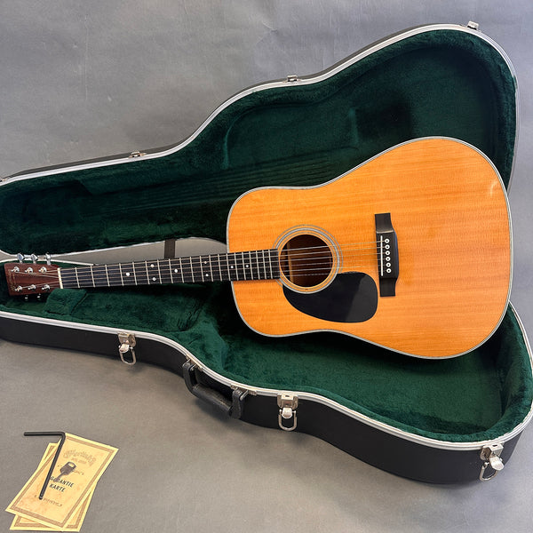 Acoustic dreadnought guitar with natural wood finish resting in open black hard shell case with green plush interior, accompanied by warranty cards and adjustment tool on gray surface