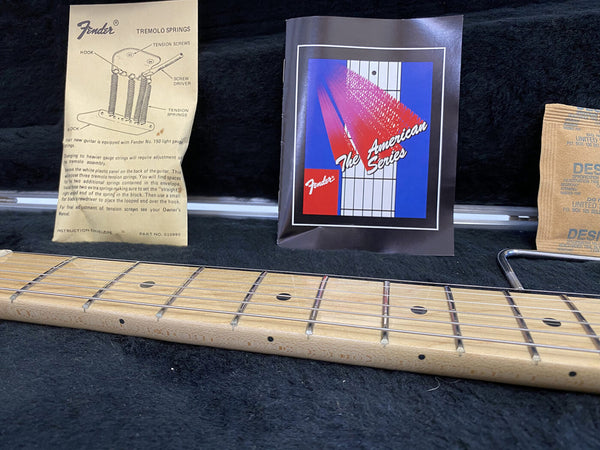 Close-up of maple guitar neck and fretboard with Fender Tremolo Springs instruction sheet, Fender The American Series booklet, and tremolo arm on black fabric background