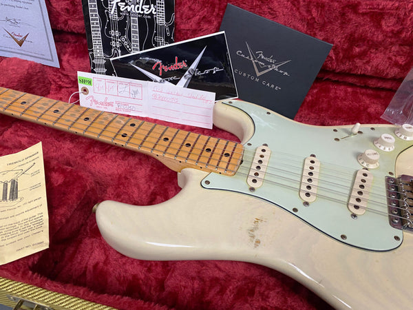 Close-up of Fender Stratocaster electric guitar with maple fretboard and vintage white body resting in red plush guitar case, accompanied by Fender Custom Shop care booklet, tremolo springs adjustment guide, and Fender paperwork.