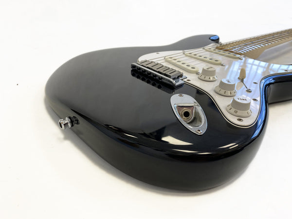 Close-up of black electric guitar body with white pickguard, tone and volume control knobs, input jack, and metal bridge on white background
