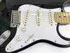 Close-up of black electric guitar body with white pickguard, three single-coil pickups, chrome bridge, volume and tone control knobs, and maple neck fingerboard