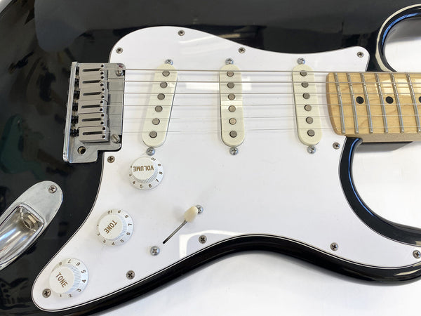Close-up of black electric guitar body with white pickguard, three single-coil pickups, chrome bridge, volume and tone control knobs, and maple neck fingerboard