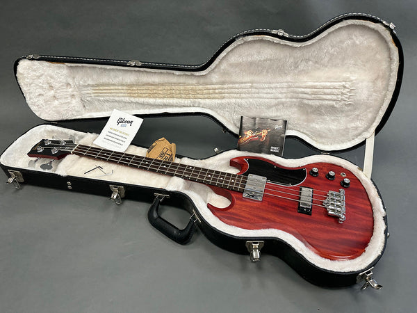 Gibson electric bass guitar with dark wood finish in open black hardshell case with plush white interior, Gibson owner's manual and accessories displayed