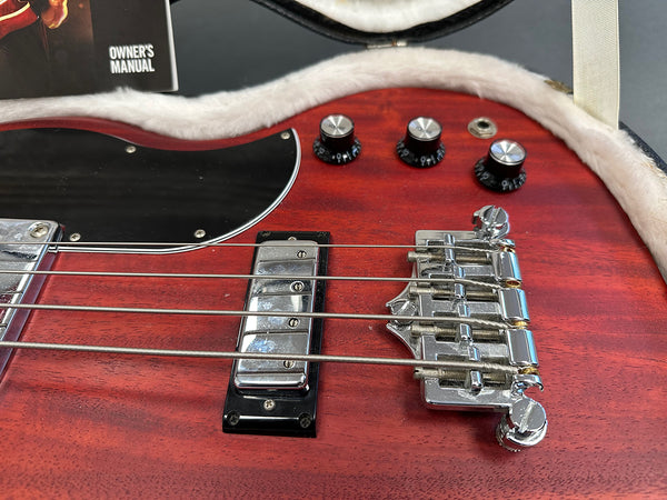 Close-up of red electric bass guitar body showing chrome bridge, pickup, black pickguard, and control knobs with an owner's manual in soft case lining