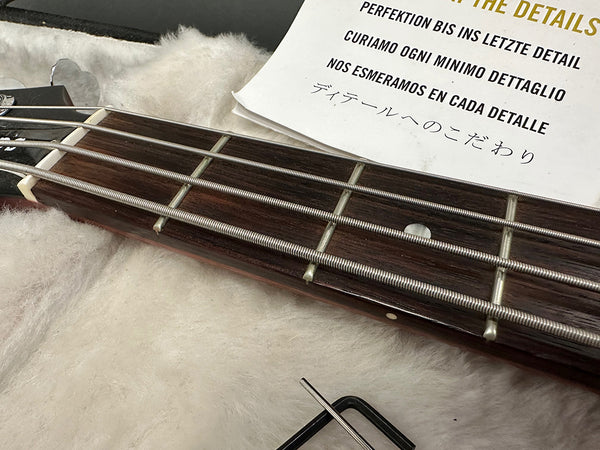 Close-up of bass guitar fretboard with metal strings, tuning peg, and a note highlighting attention to detail in multiple languages on soft fabric background, including Allen wrench tool for guitar setup.