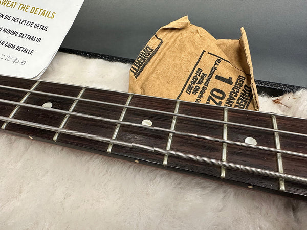 Close-up of bass guitar fretboard with metal strings and dot inlays, showing detailed wood grain and a desiccant packet in the background inside a guitar case lining