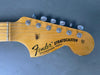 Close-up of Fender Stratocaster guitar headstock with six tuning pegs, natural wood finish, and "Fender Stratocaster with Synchronized Tremolo" logo on the front against gray background.