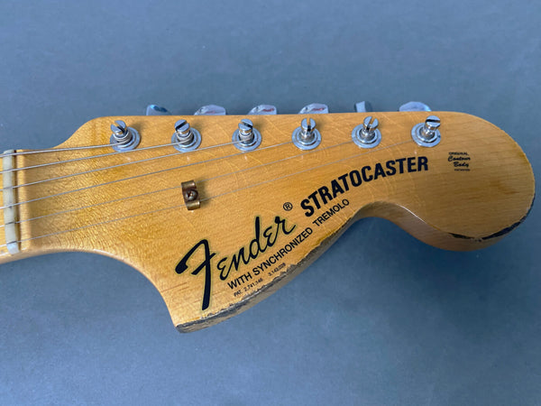 Close-up of Fender Stratocaster guitar headstock with six tuning pegs, natural wood finish, and black Fender logo on a gray background.
