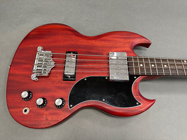 Close-up of red electric bass guitar body with chrome bridge, two pickups, black pickguard, three black control knobs, and four strings on gray background