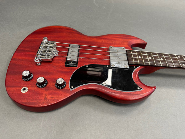 Close-up view of red electric bass guitar body with black pickguard, chrome bridge and pickups, and three control knobs on gray background