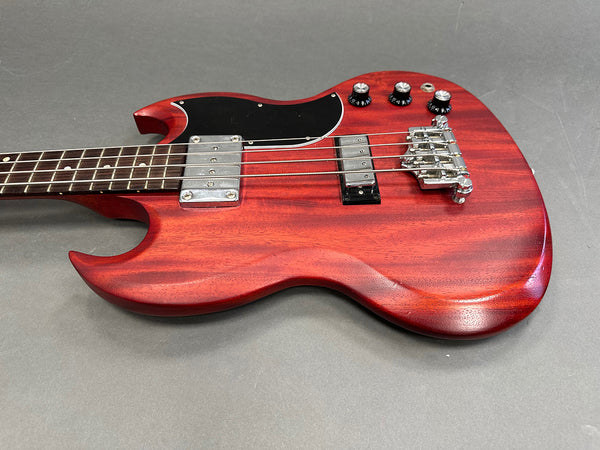 Close-up of red electric bass guitar body with black pickguard, chrome pickups, bridge, and control knobs on gray background