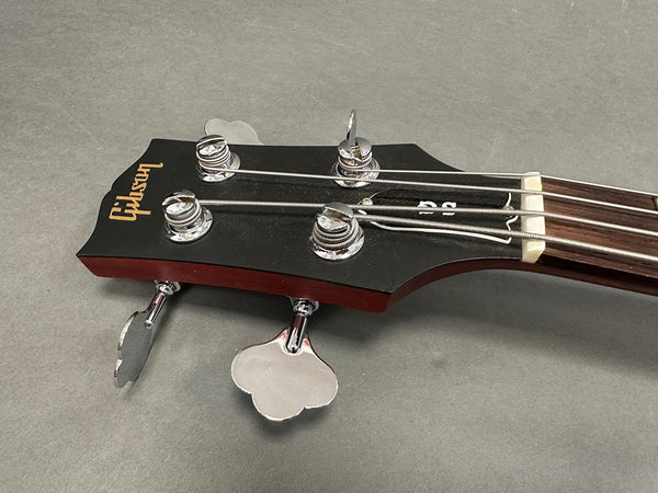 Close-up image of Gibson bass guitar headstock with chrome tuning pegs and strings on gray background