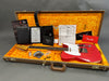 Fender vintage red electric guitar with white pickguard in brown hardshell case lined with orange plush, displaying Fender certificate of authenticity, owner's manual, custom shop care booklet, guitar strap, pick, and bridge cover plate accessories.