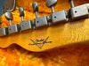 Close-up of vintage Fender Custom Shop guitar headstock with tuning pegs and logo on figured maple wood background in plush yellow case lining.
