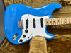 Close-up of bright blue electric guitar with white pickguard and maple neck resting on brown plush guitar case interior