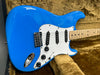 Close-up of blue electric guitar body with white pickguard, three single-coil pickups, volume and tone control knobs, and maple neck on plush brown case lining