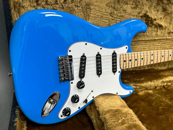 Close-up of blue electric guitar body with white pickguard, three single-coil pickups, volume and tone control knobs, and maple neck on plush brown case lining