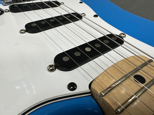 Close-up of blue electric guitar body showing white pickguard, black single-coil pickups, metal strings, and natural wood fretboard.