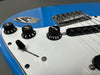 Close-up of blue electric guitar body showing white pickguard, black tone and volume control knobs, pickup, and chrome bridge with strings.