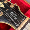 Close-up of a vintage black guitar headstock with "Lola Paul Custom" logo, metal tuning pegs, and nylon strings inside a red velvet-lined guitar case