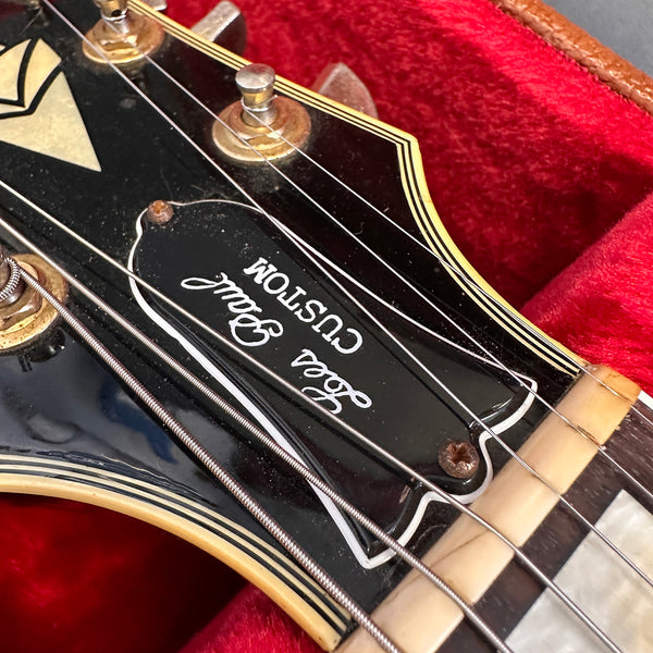 Close-up of a vintage black guitar headstock with "Lola Paul Custom" logo, metal tuning pegs, and nylon strings inside a red velvet-lined guitar case