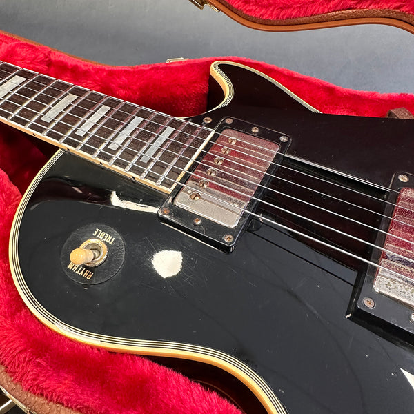 Close-up of black electric guitar body with chrome pickups and rhythm/treble toggle switch, resting in red plush-lined guitar case.