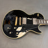 Black solid body electric guitar with dual humbucker pickups, gold hardware, and four black control knobs on gray background