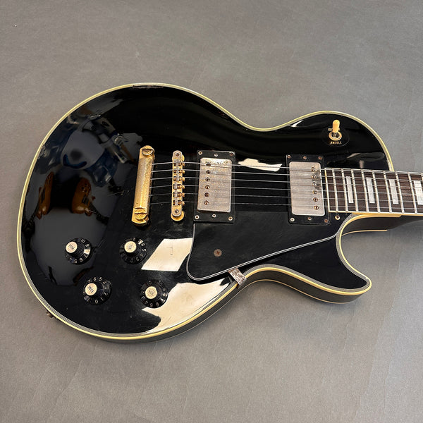 Black solid body electric guitar with dual humbucker pickups, gold hardware, and four black control knobs on gray background