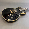 Black electric guitar with cream binding and gold hardware featuring four black control knobs labeled tone and volume on gray surface