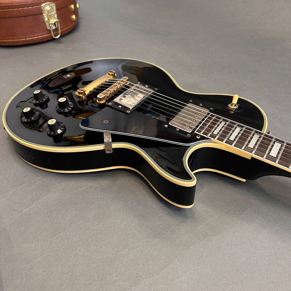 Black electric guitar with gold hardware and multiple control knobs on a gray surface with a leather guitar case visible in the background