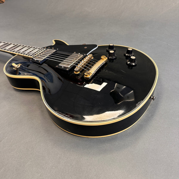 Close-up of black electric guitar with gold hardware, four black control knobs, and cream binding on the body and neck, laid on gray surface.