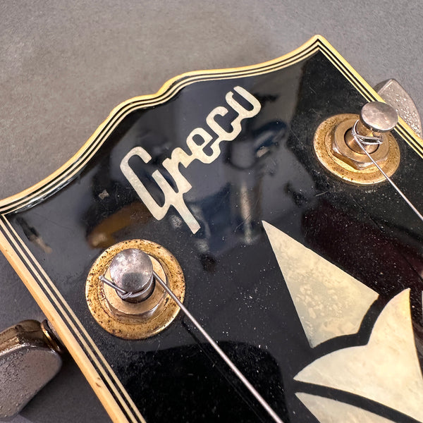 Close-up of vintage Greco guitar headstock showing tuning pegs, logo, and decorative inlay on black background