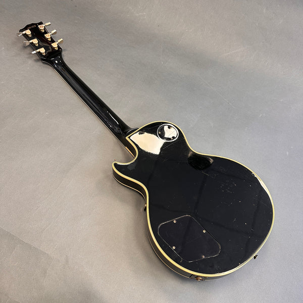 Black solid body electric guitar with cream binding and six tuning pegs, back view on gray surface, showing scratches and wear