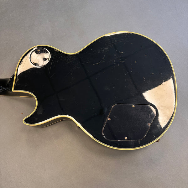 Back of black electric guitar with cream binding and visible wear and scratches, showing control cavity covers on gray background