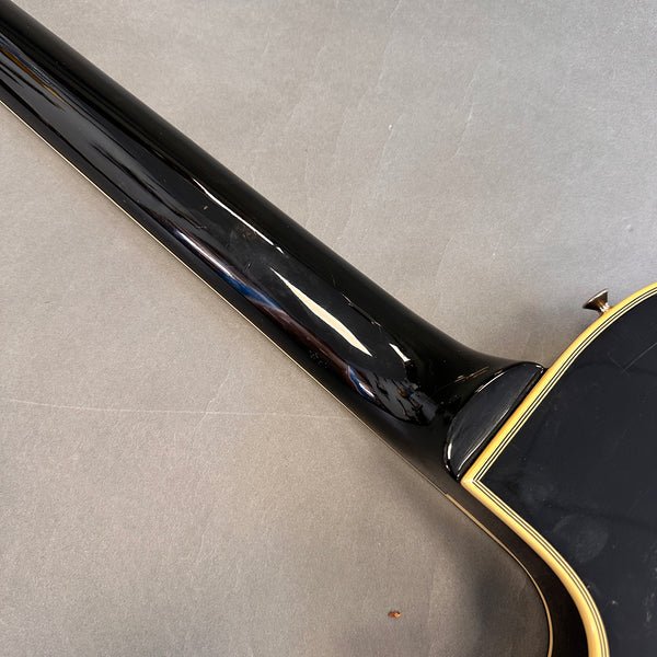 Close-up of the neck and heel joint of a black acoustic guitar with cream binding detail on the body edge, displayed on a gray surface.