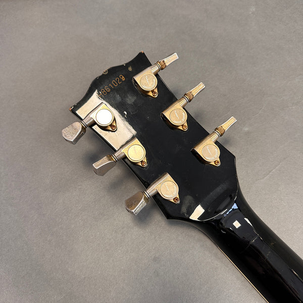 Back of black guitar headstock with six vintage gold tuning pegs labeled "Greco Guitar" and serial number 1861029 on gray background