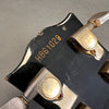 Close-up of black guitar headstock with serial number H861028 and vintage gold tuning pegs engraved with Greco Guitar logo