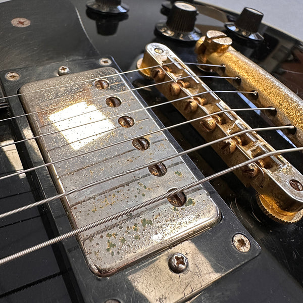 Close-up of vintage electric guitar pickup and bridge showing aged metal with rust and corrosion, black guitar body with control knobs and screws, detailed guitar strings and hardware.
