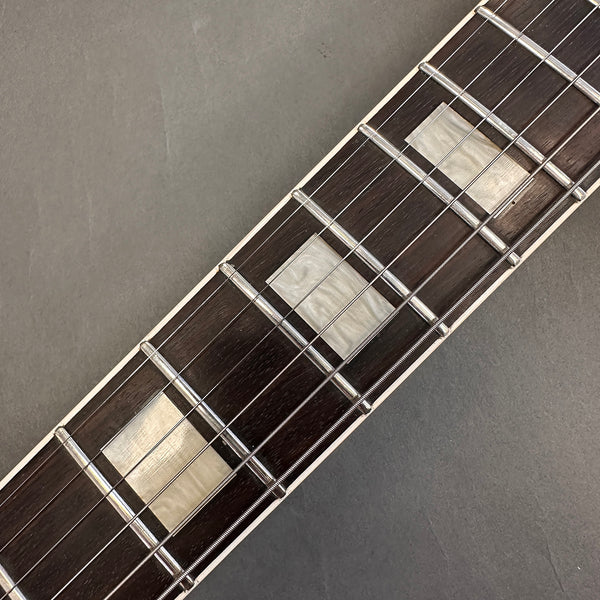 Close-up of guitar fretboard with rectangular pearl inlays and steel strings on dark wood fingerboard against gray background
