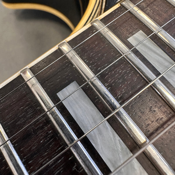 Close-up of dark wood guitar fretboard with rectangular mother-of-pearl inlays and metal frets, showing guitar strings and binding.
