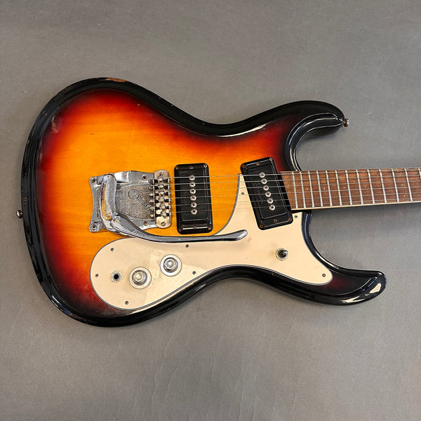 Vintage sunburst electric guitar with double cutaway body, two black single-coil pickups, white pickguard, and tremolo arm on gray background.