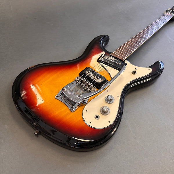 Vintage electric guitar with sunburst finish, dual pickups, white pickguard, and vibrato tailpiece on gray surface